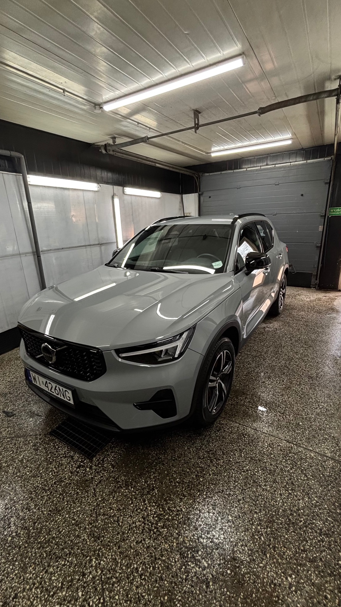 Szary samochód Volvo XC40 po umyciu, stojący wewnątrz myjni z mokrą podłogą i otwartą bramą garażową w tle.