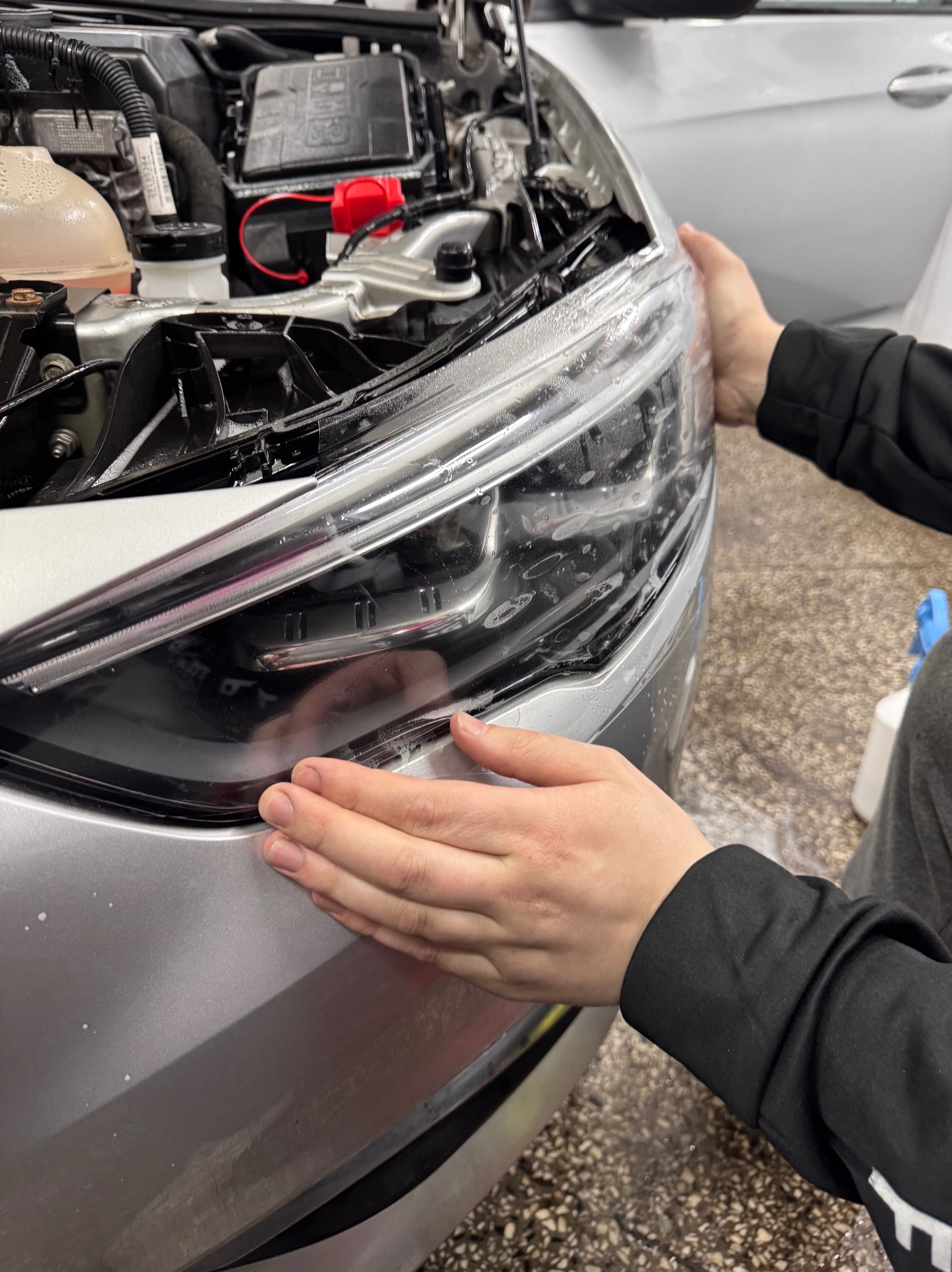 Nakładanie folii ochronnej na przedni reflektor srebrnego samochodu, widoczne dłonie pracownika wygładzające folię, z bliska, z otwartą maską.