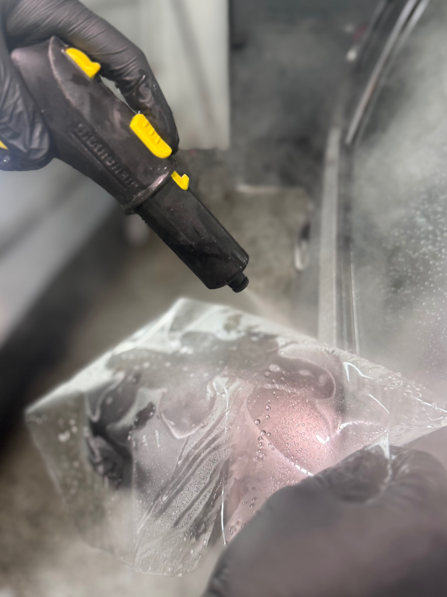 Czyszczenie parowe elementu tapicerki samochodowej przez osobę w rękawicach, z użyciem parownicy z żółtymi akcentami, w pomieszczeniu z metalowymi elementami.