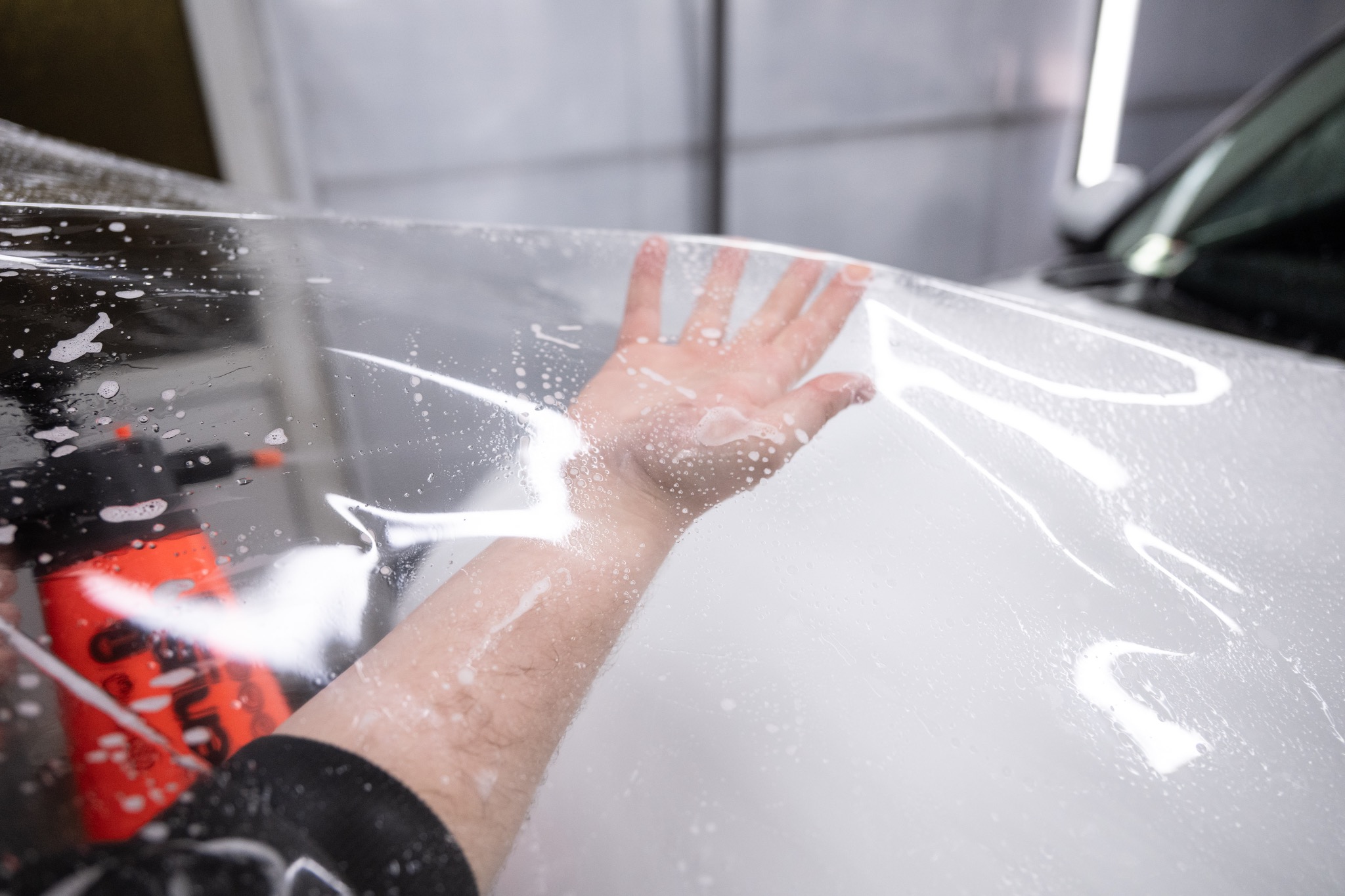 Nakładanie folii ochronnej na maskę białego samochodu w warsztacie. Widoczna ręka pracownika i spryskiwacz. Oświetlenie górne.