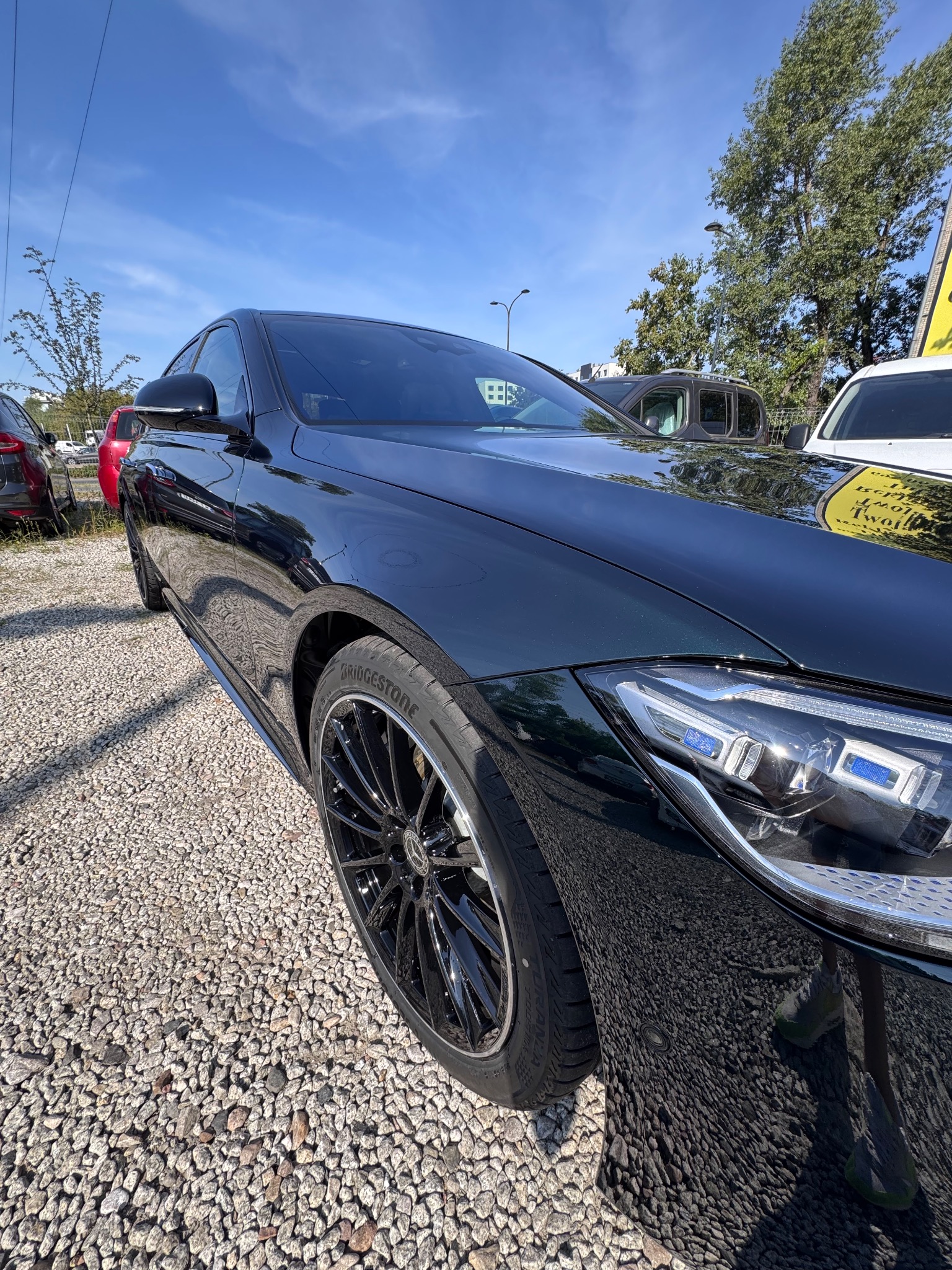 Błyszczący, ciemnozielony samochód z czarnymi felgami Bridgestone, zaparkowany na żwirowym parkingu w słoneczny dzień. Widoczny fragment reflektora i karoserii.