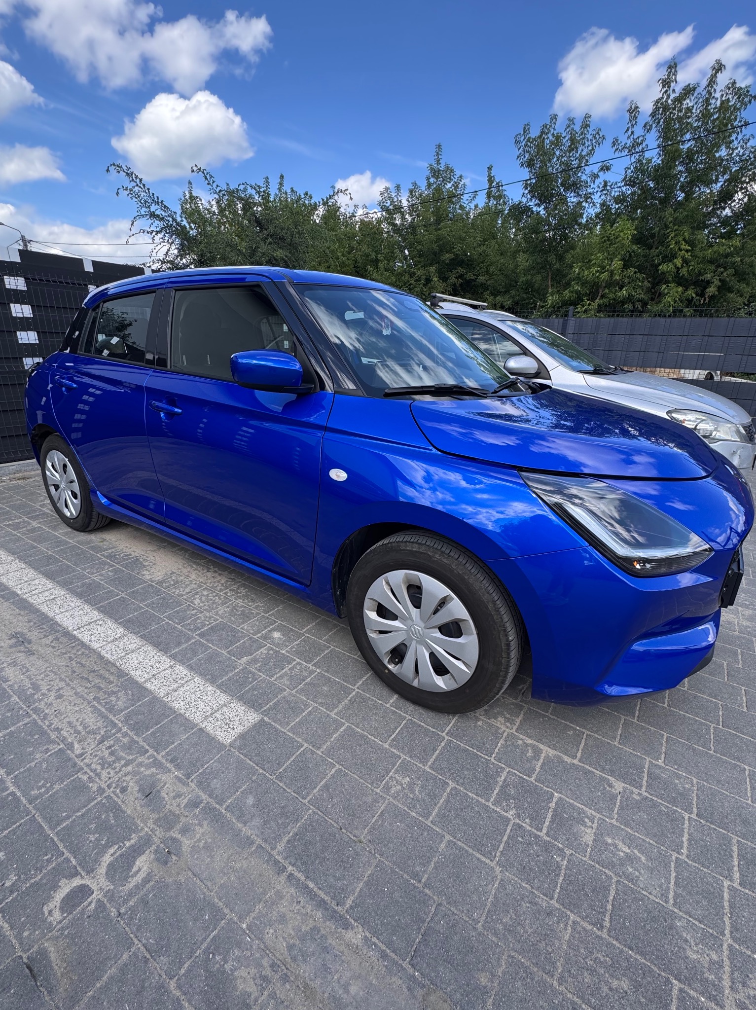 Błyszczący, granatowy samochód na kostce brukowej, z widocznym srebrnym autem w tle i błękitnym niebem z chmurami. Auto po detailingu, z wyraźnym odbiciem światła.