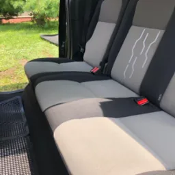 Wnętrze samochodu z widoczną tylną kanapą w odcieniach szarości i czerni, z detalami w postaci czerwonych klamer pasów bezpieczeństwa i czarnych gumowych dywaników. Trawa widoczna przez otwarte drzwi.