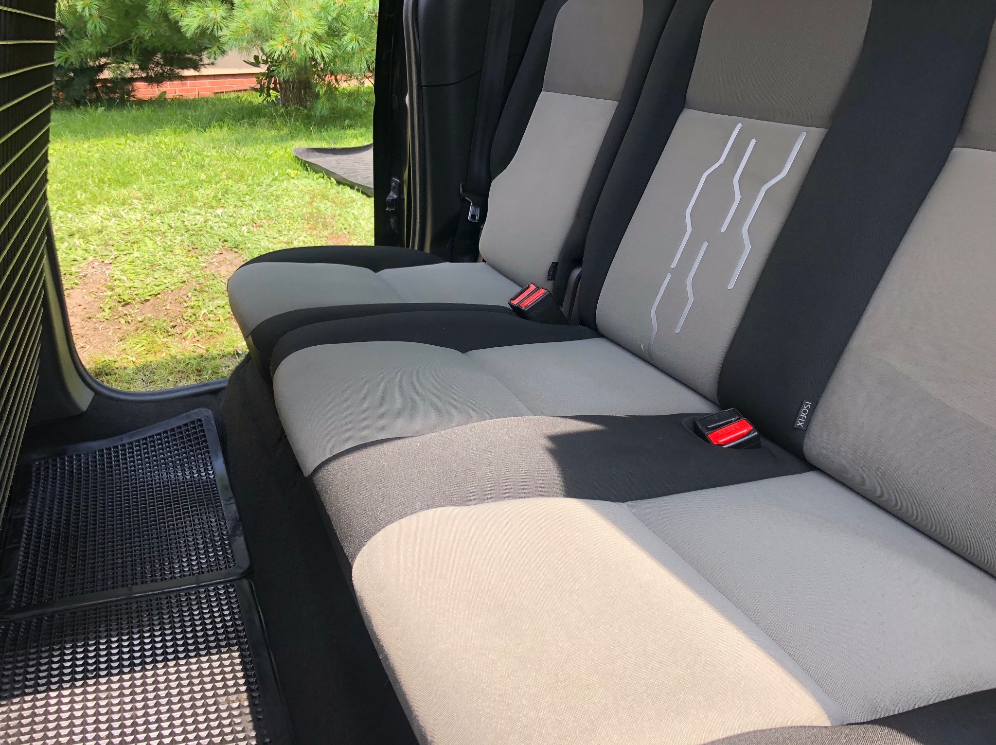 Wnętrze samochodu z widoczną tylną kanapą w odcieniach szarości i czerni, z detalami w postaci czerwonych klamer pasów bezpieczeństwa i czarnych gumowych dywaników. Trawa widoczna przez otwarte drzwi.