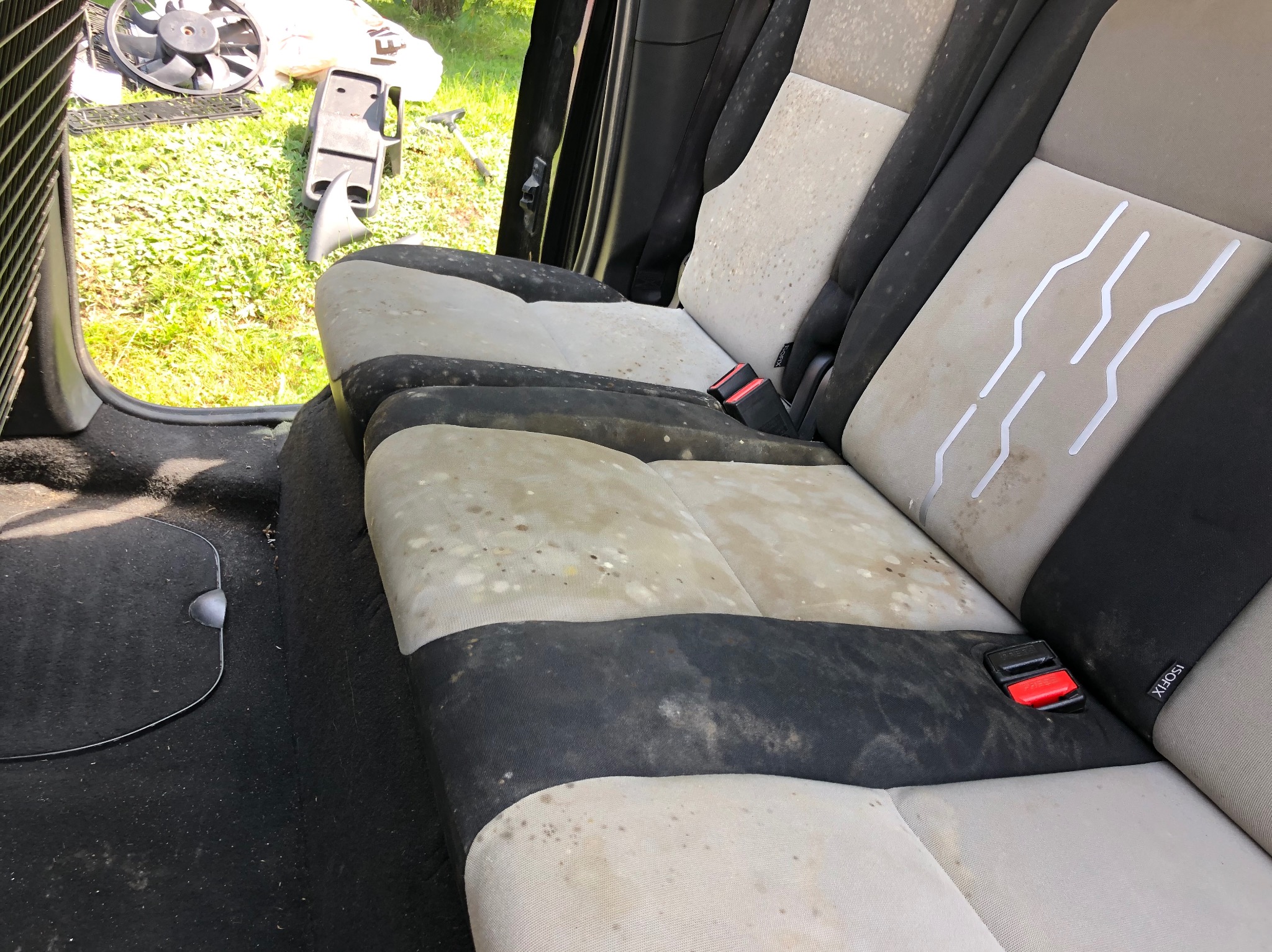 Bardzo zabrudzone, jasne siedzenia samochodowe z czarnymi wstawkami i czerwonymi klamrami pasów bezpieczeństwa, widoczne plamy i zacieki na tapicerce.