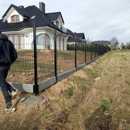 MT Ogrodzenia Marek Tymecki - Nowoczesne ogrodzenie panelowe z podmurówką betonową, czarne panele na tle białego domu z ciemnym dachem, widoczny fragment osoby podczas montażu.