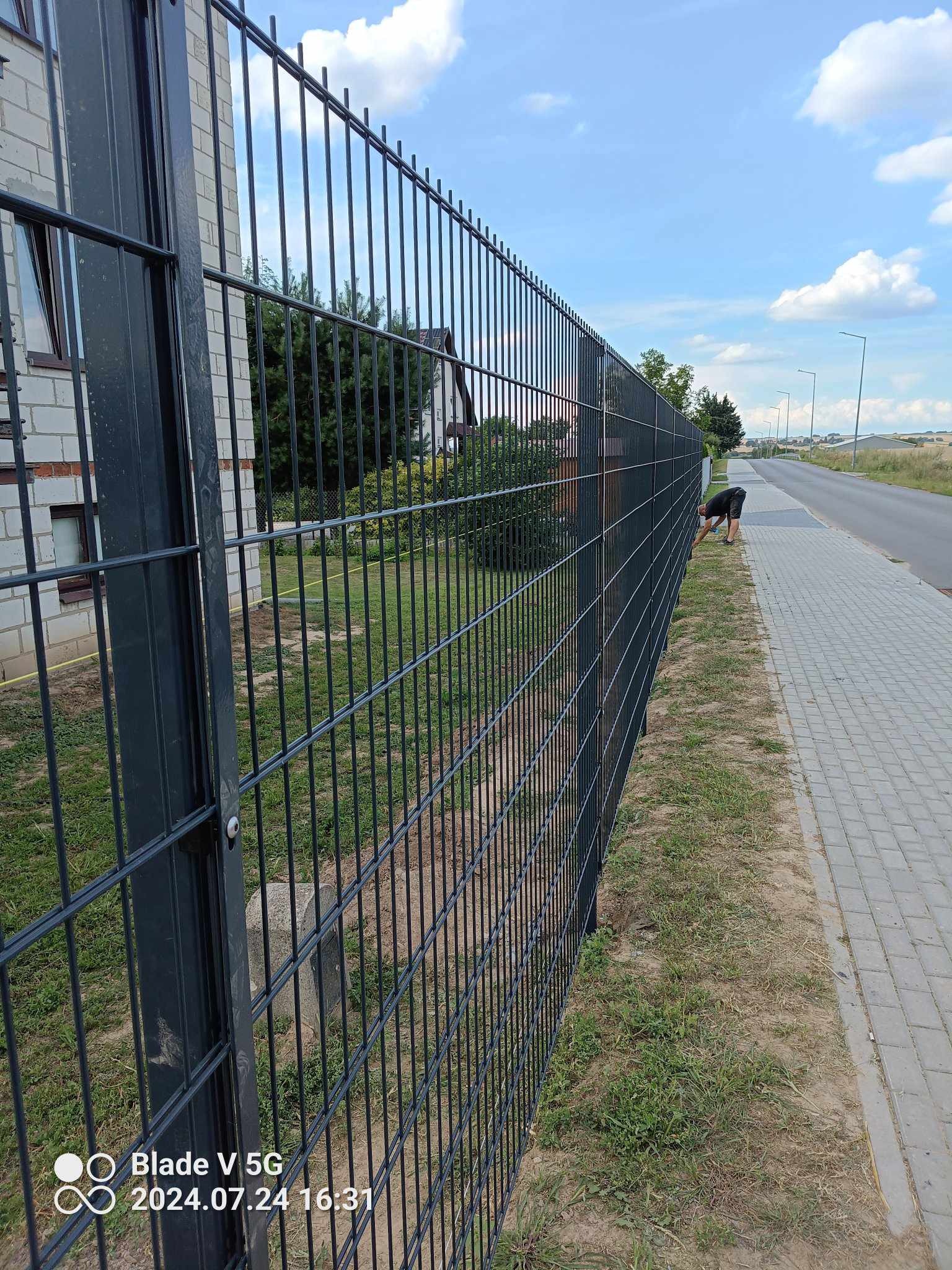 Nowoczesne ogrodzenie panelowe w kolorze grafitowym, oddzielające posesję od chodnika i drogi. Widoczny fragment budynku z jasnej cegły i osoba przy krawędzi ogrodzenia.