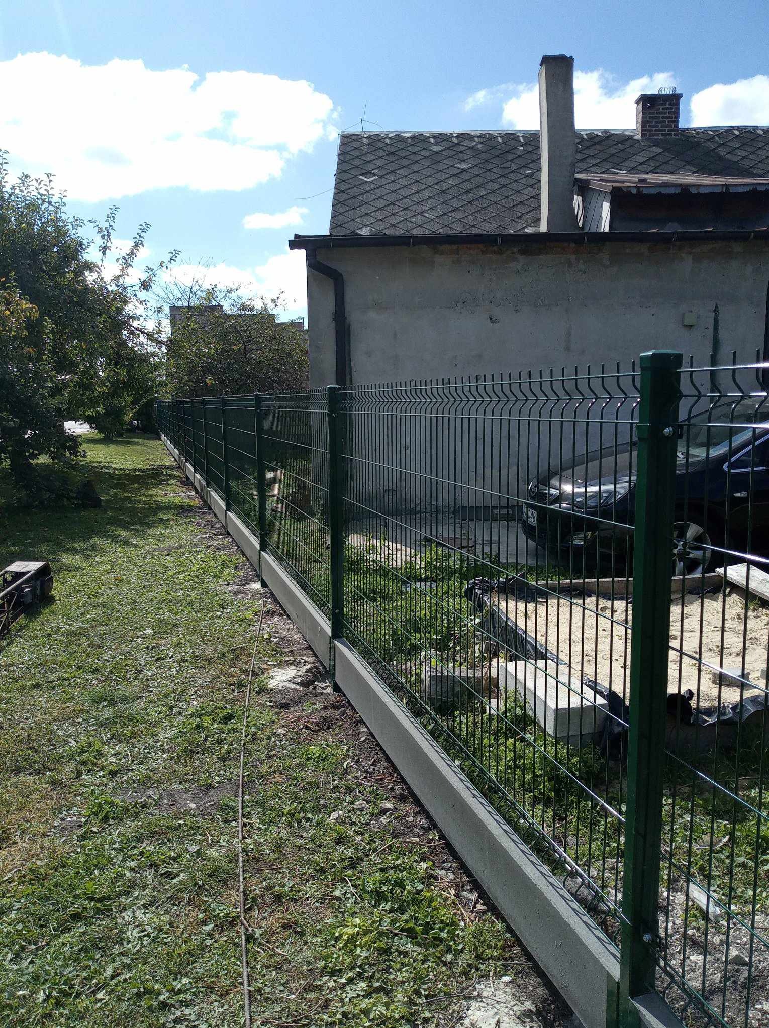 Długi, zielony panelowy płot z betonową podmurówką, biegnący wzdłuż posesji. W tle dom z szarym dachem i zaparkowany samochód. Słoneczny dzień.