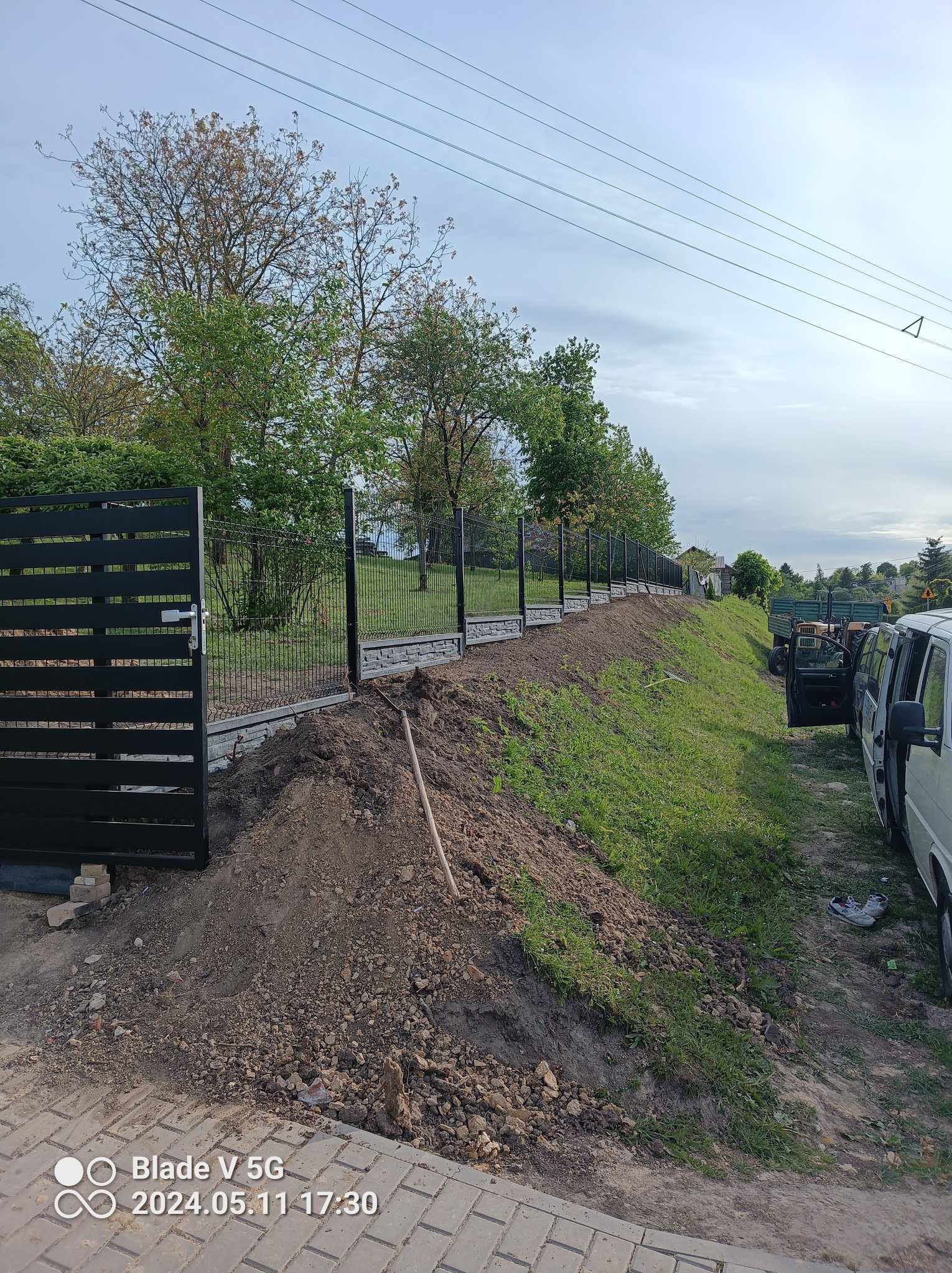 Montaż nowoczesnego ogrodzenia panelowego z elementami betonowymi na nierównym terenie. Widoczna brama wjazdowa i częściowo ułożona kostka brukowa.