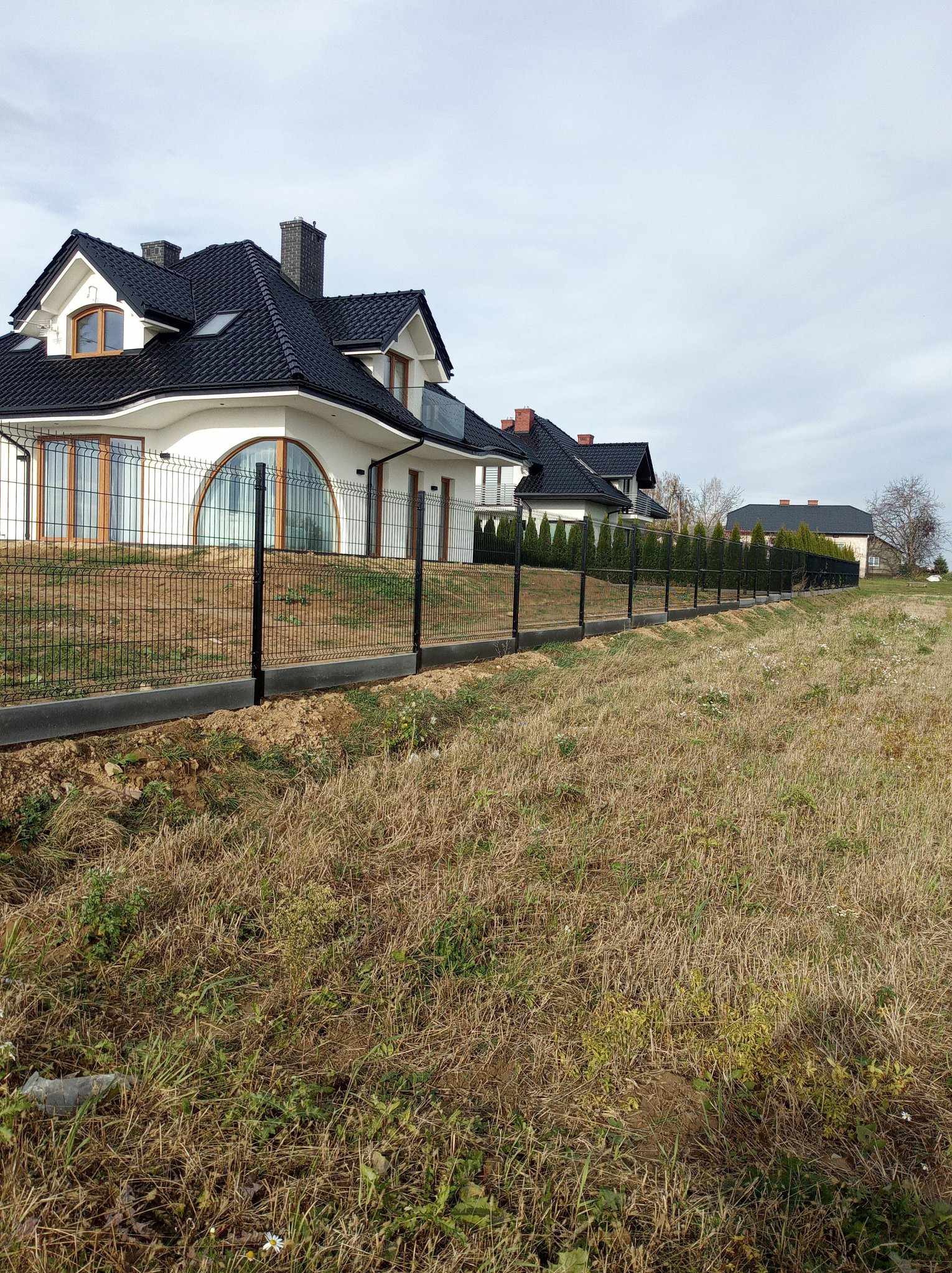 Długa linia czarnego ogrodzenia panelowego z betonową podmurówką, oddzielająca posesję z nowoczesnym domem od zarośniętego pola. Widok z perspektywy ziemi.