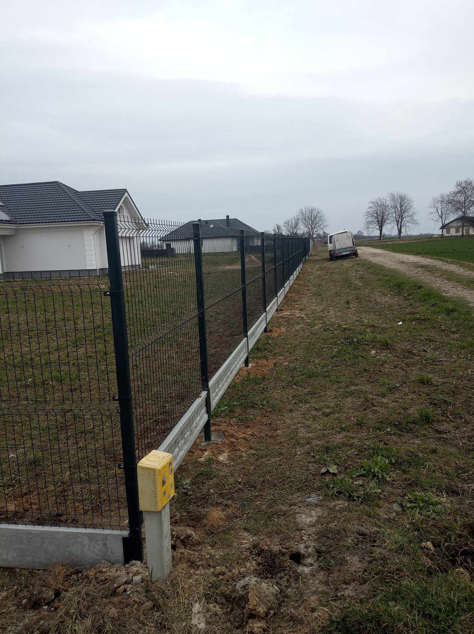 Nowoczesne ogrodzenie panelowe z podmurówką betonową, widok z boku, z żółtą skrzynką gazową na słupku. W tle domy i polna droga z białym busem.