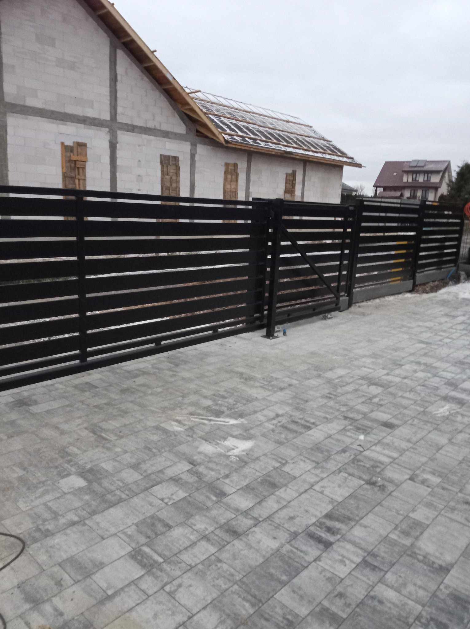 Nowoczesne, poziome, czarne ogrodzenie metalowe z przesuwną bramą na tle szarej kostki brukowej i budynku w trakcie budowy w pochmurny dzień.