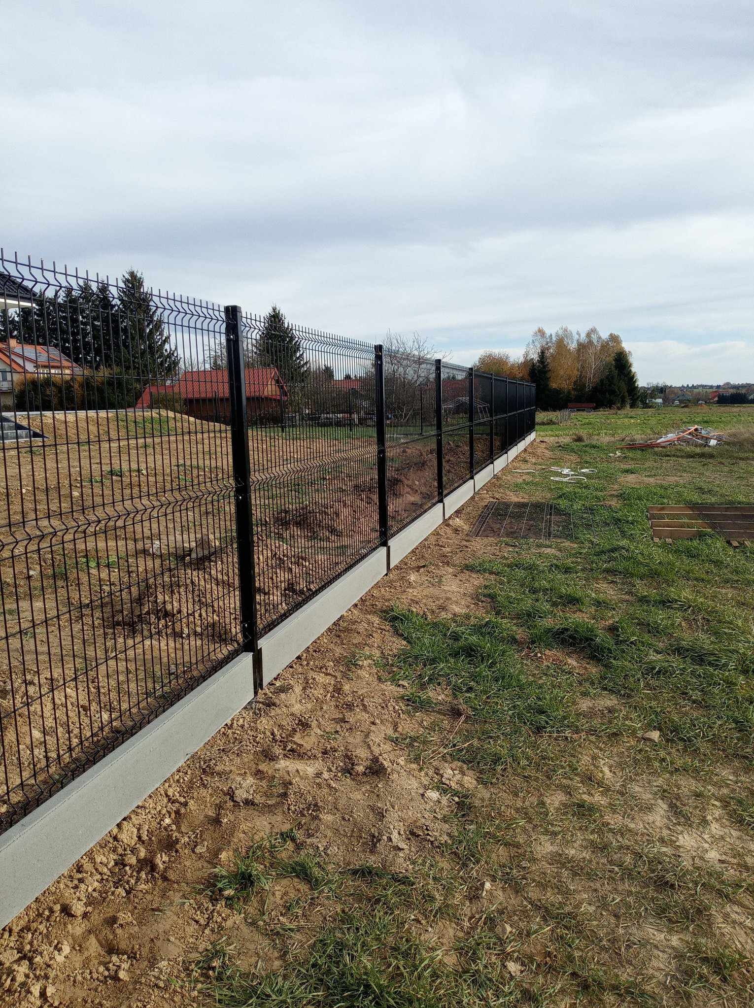 Czarny panelowy płot z podmurówką na nierównym terenie, widok z boku, w tle domy i drzewa. Charakterystyczne łączenie paneli na słupkach.