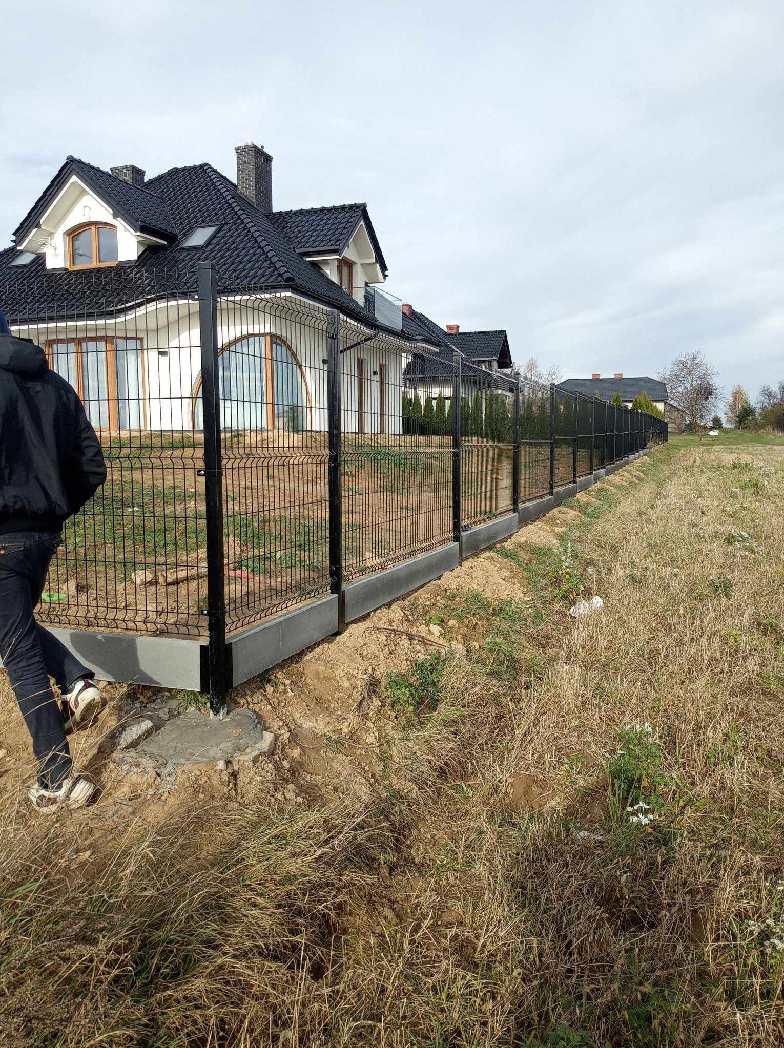 Nowoczesne ogrodzenie panelowe z podmurówką betonową, czarne panele na tle białego domu z ciemnym dachem, widoczny fragment osoby podczas montażu.
