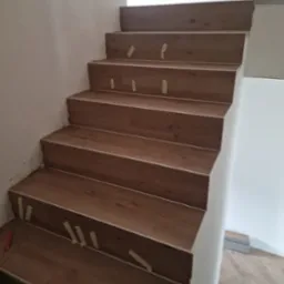 Schody wyłożone panelami imitującymi drewno, zabezpieczone taśmą malarską na czas montażu, widoczne panele podłogowe w tle.