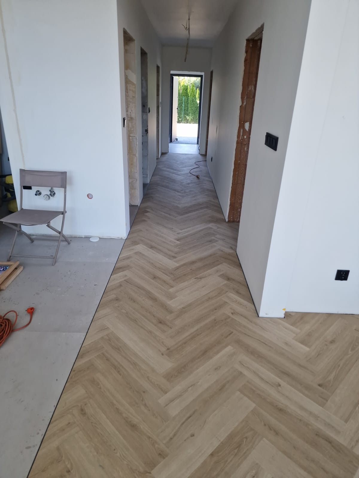 Korytarz w trakcie remontu w Chorzowie z nowo ułożonym parkietem w jodełkę, widoczny fragment podłogi przed położeniem parkietu, krzesło i kable.