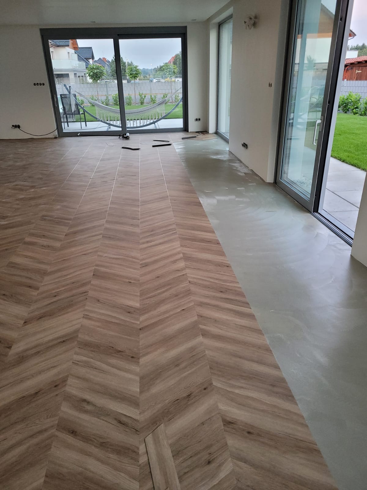 Fragment podłogi w trakcie układania, widoczny wzór jodełki z paneli drewnopodobnych, kontrastujący z surowym betonem w tle, duże okna przesuwne wychodzące na ogród.