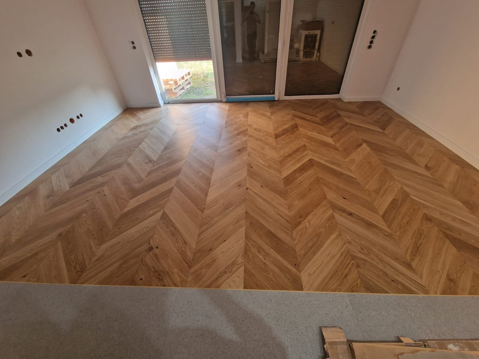 Ukończona podłoga z parkietu ułożonego we wzór jodełki francuskiej w jasnym pomieszczeniu z oknami balkonowymi i szarym progiem.
