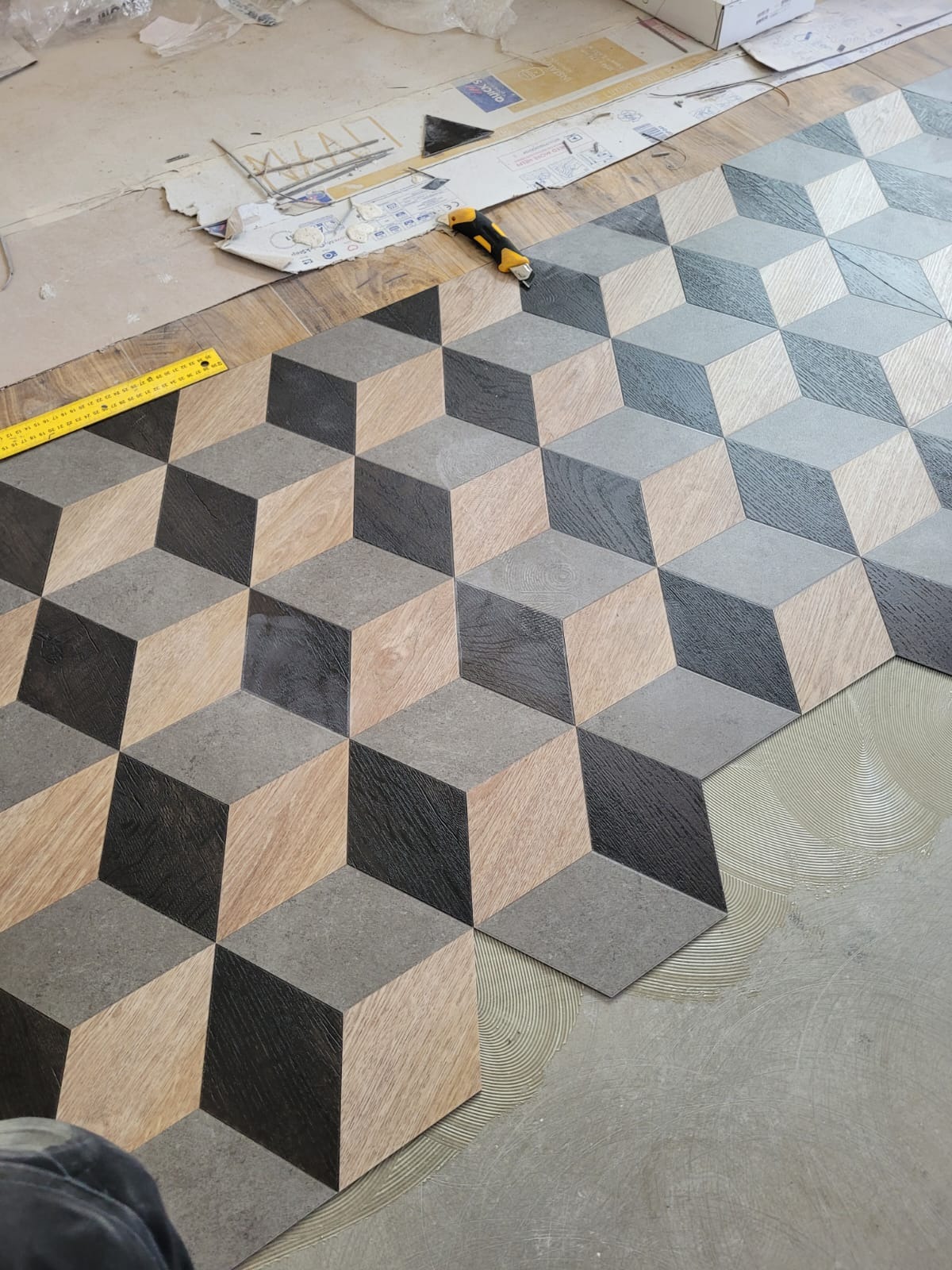 Układanie podłogi z paneli o wzorze 3D: widoczny fragment ułożonej podłogi z efektem trójwymiarowych kostek w odcieniach szarości i drewna, obok żółta miarka, nóż do cięcia paneli i resztki...