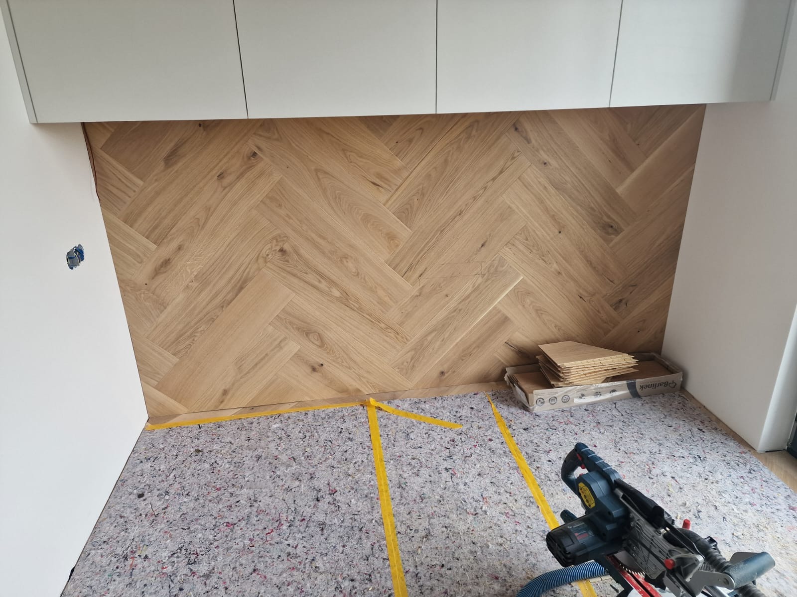 Fragment pokoju z ułożonym parkietem w jodełkę francuską, widoczna maszyna do cięcia parkietu, podkład wygłuszający i taśma malarska.