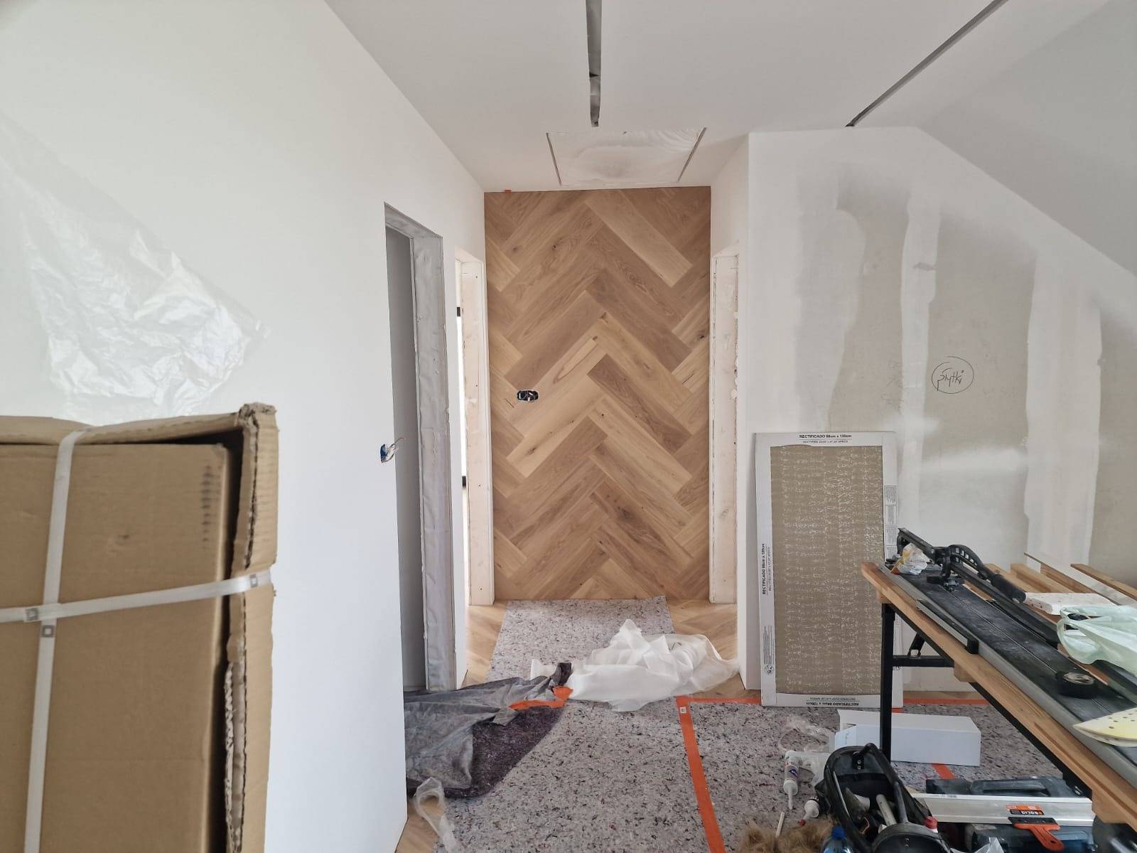 Korytarz w trakcie remontu z ułożonym parkietem w jodełkę na ścianie, widoczne narzędzia i materiały budowlane, ściany w stanie surowym.