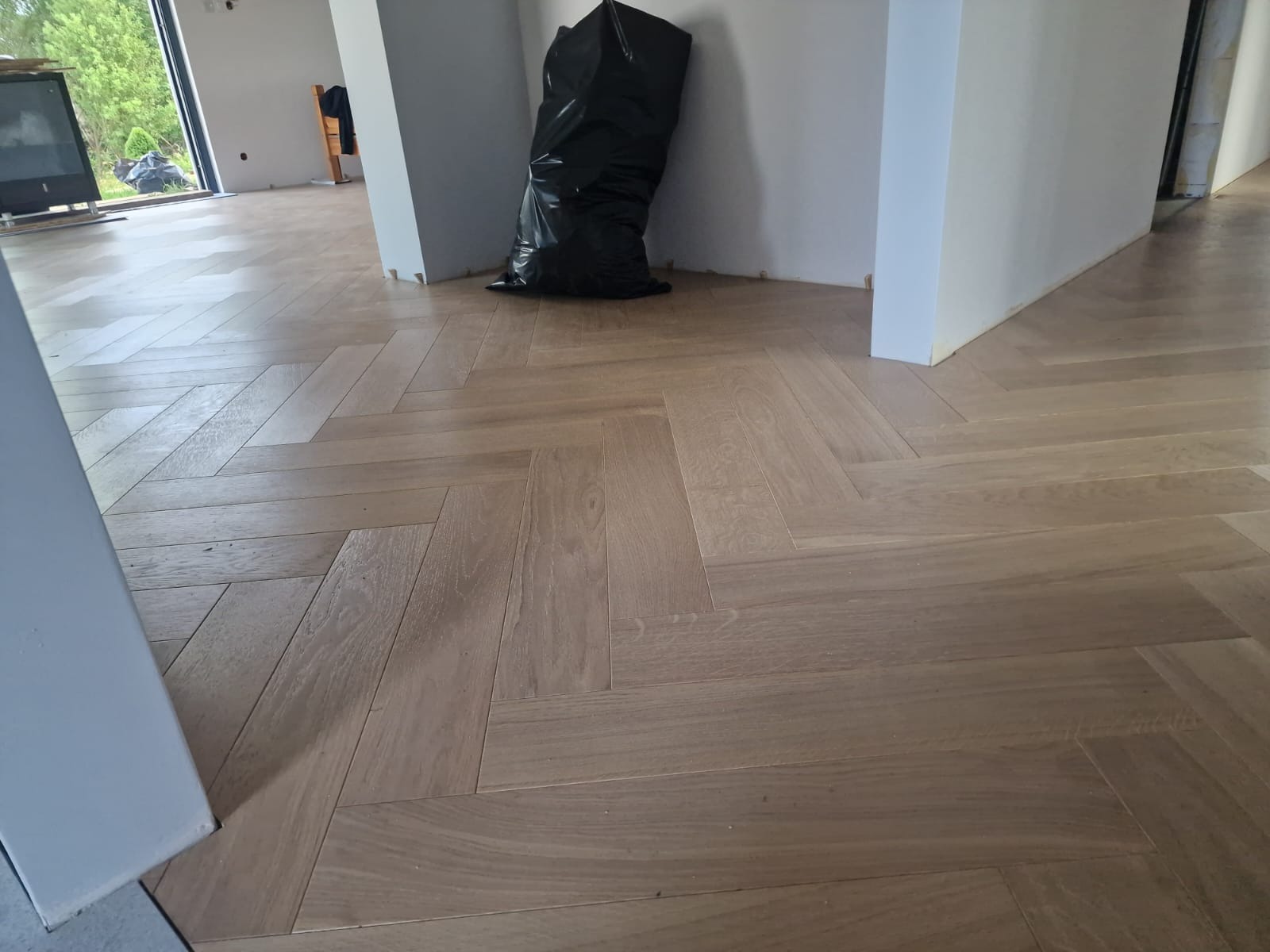 Jasny parkiet ułożony we wzór jodełki francuskiej, widoczny czarny worek na śmieci i fragmenty ścian w trakcie remontu.