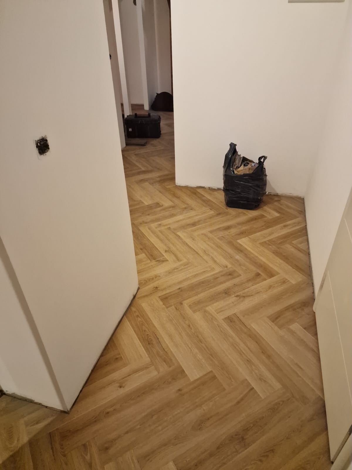 Ułożony parkiet w jodełkę w jasnym odcieniu drewna, widoczny czarny worek na śmieci i skrzynka narzędziowa w tle, ściany w trakcie wykończenia.