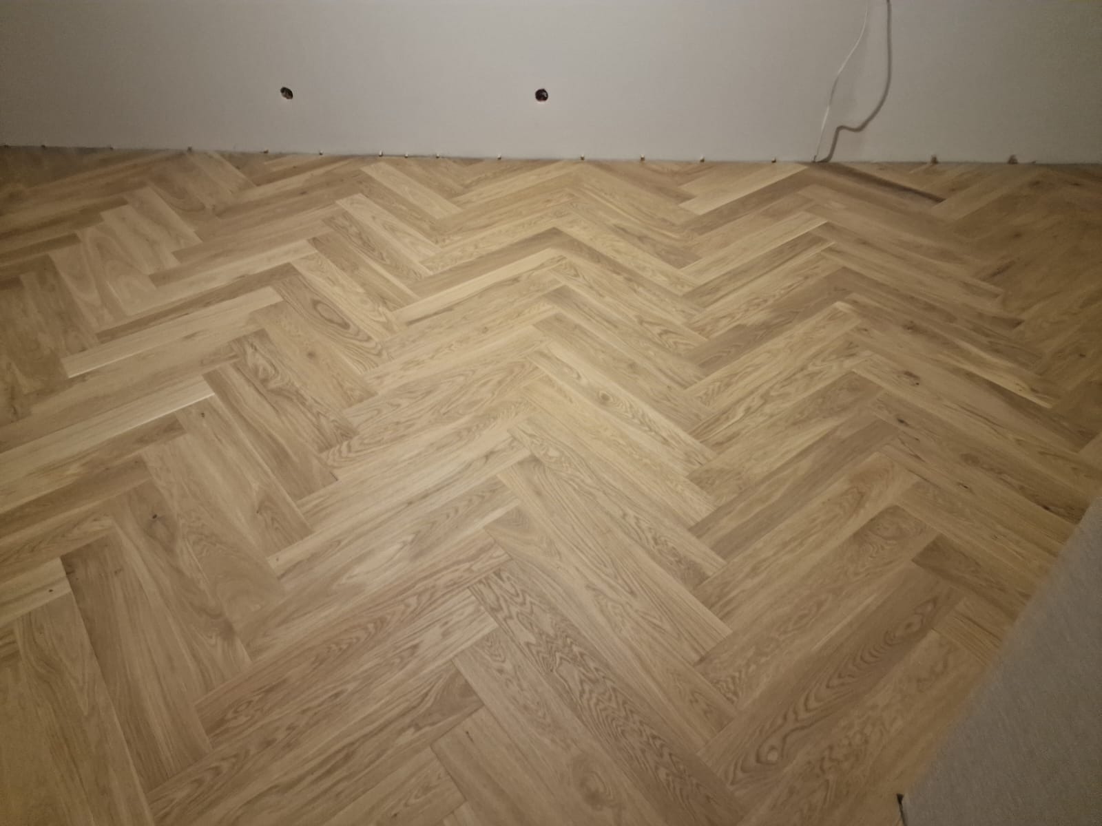 Jasny parkiet ułożony we wzór jodełki francuskiej, widoczne listwy dystansowe przy ścianie i wystające kable elektryczne.