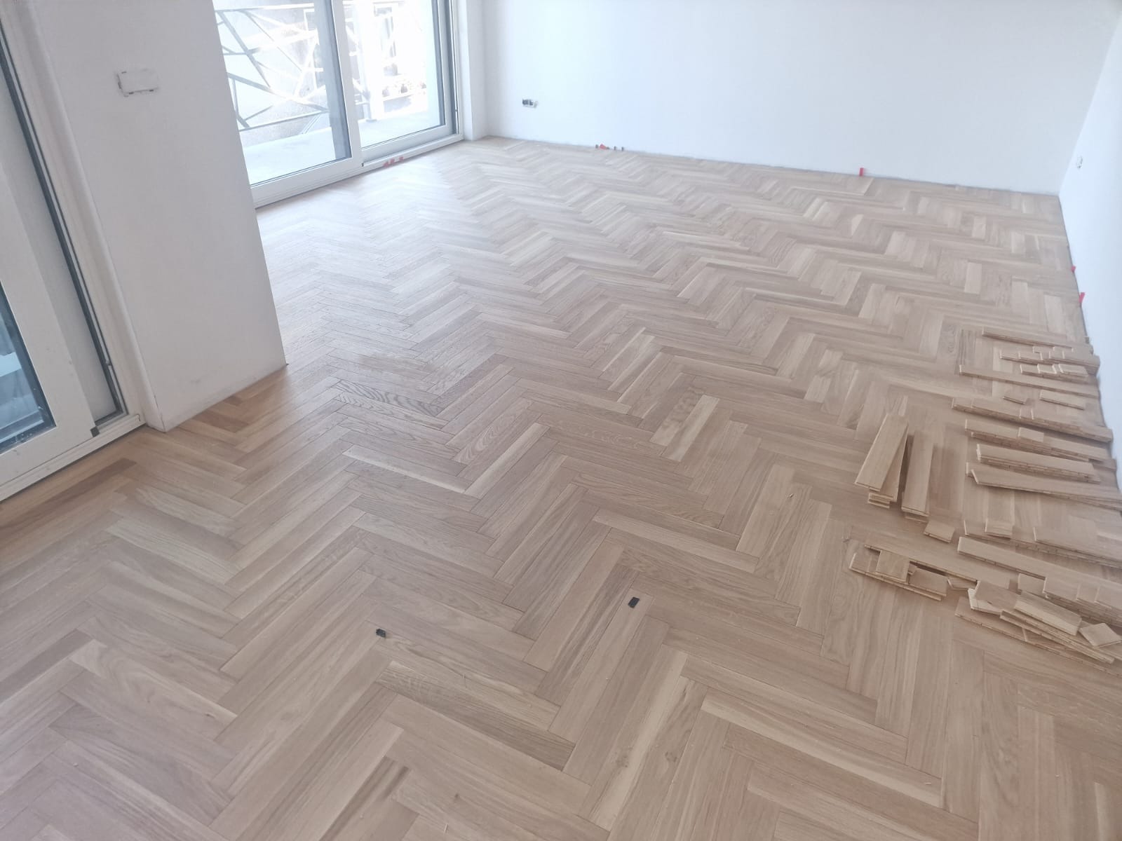 Jasny parkiet ułożony we wzór jodełki francuskiej, z widocznymi elementami dystansowymi przy ścianie i stertą nieułożonych klepek parkietowych w rogu pomieszczenia.