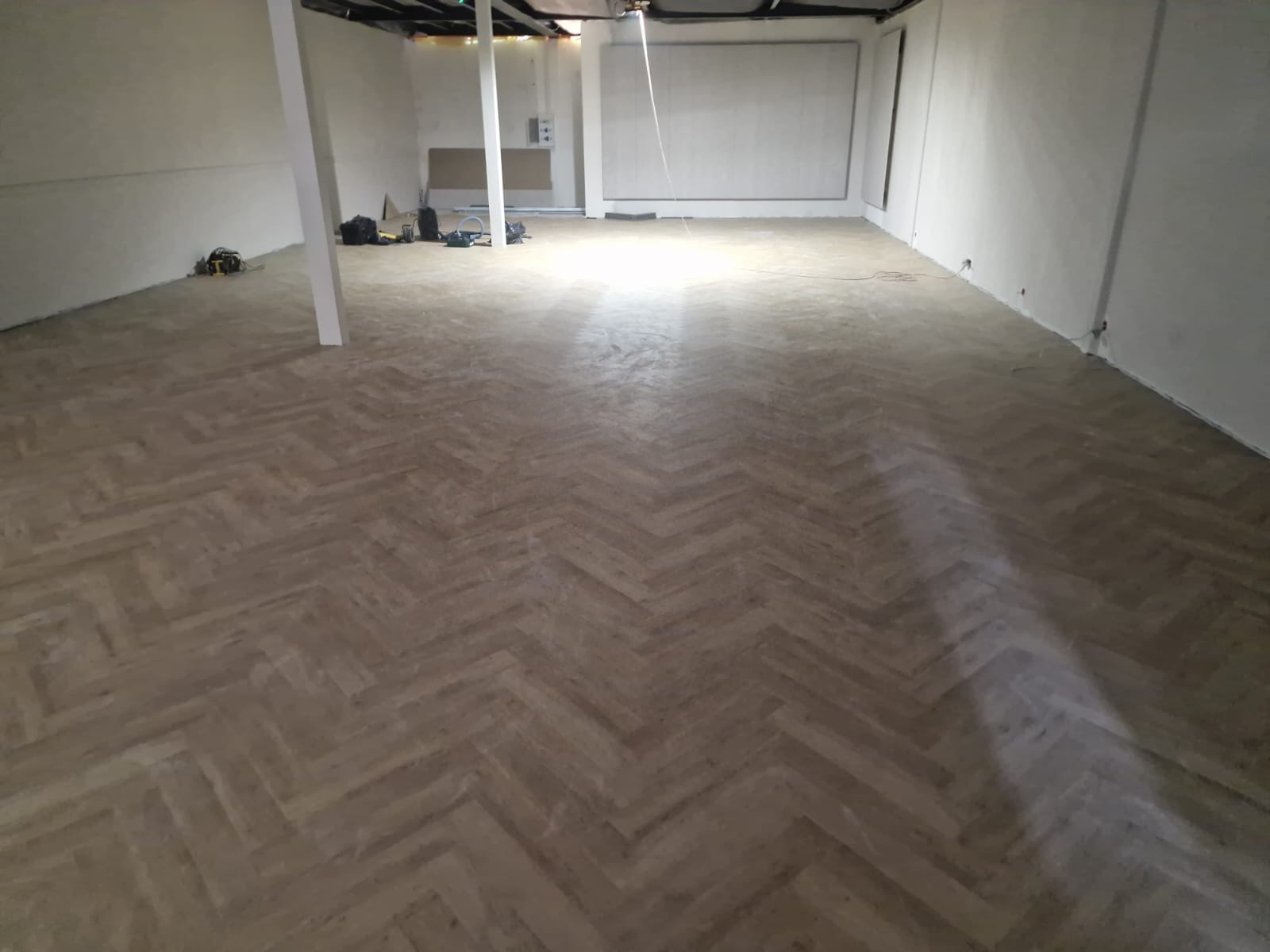 Ułożony parkiet w jodełkę w jasnym, pustym pomieszczeniu z widocznymi narzędziami w tle.