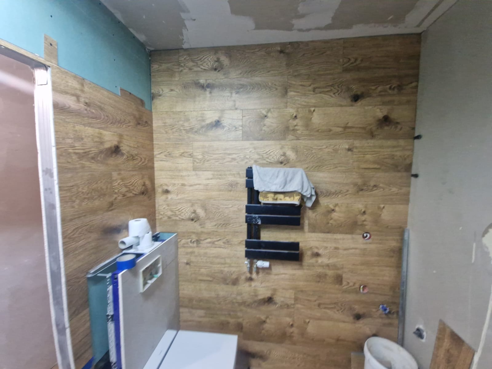 Łazienka w trakcie remontu: ściany wyłożone panelami imitującymi drewno, widoczne elementy instalacji sanitarnej i grzejnik drabinkowy z ręcznikiem, surowy stan wykończenia sufitu.