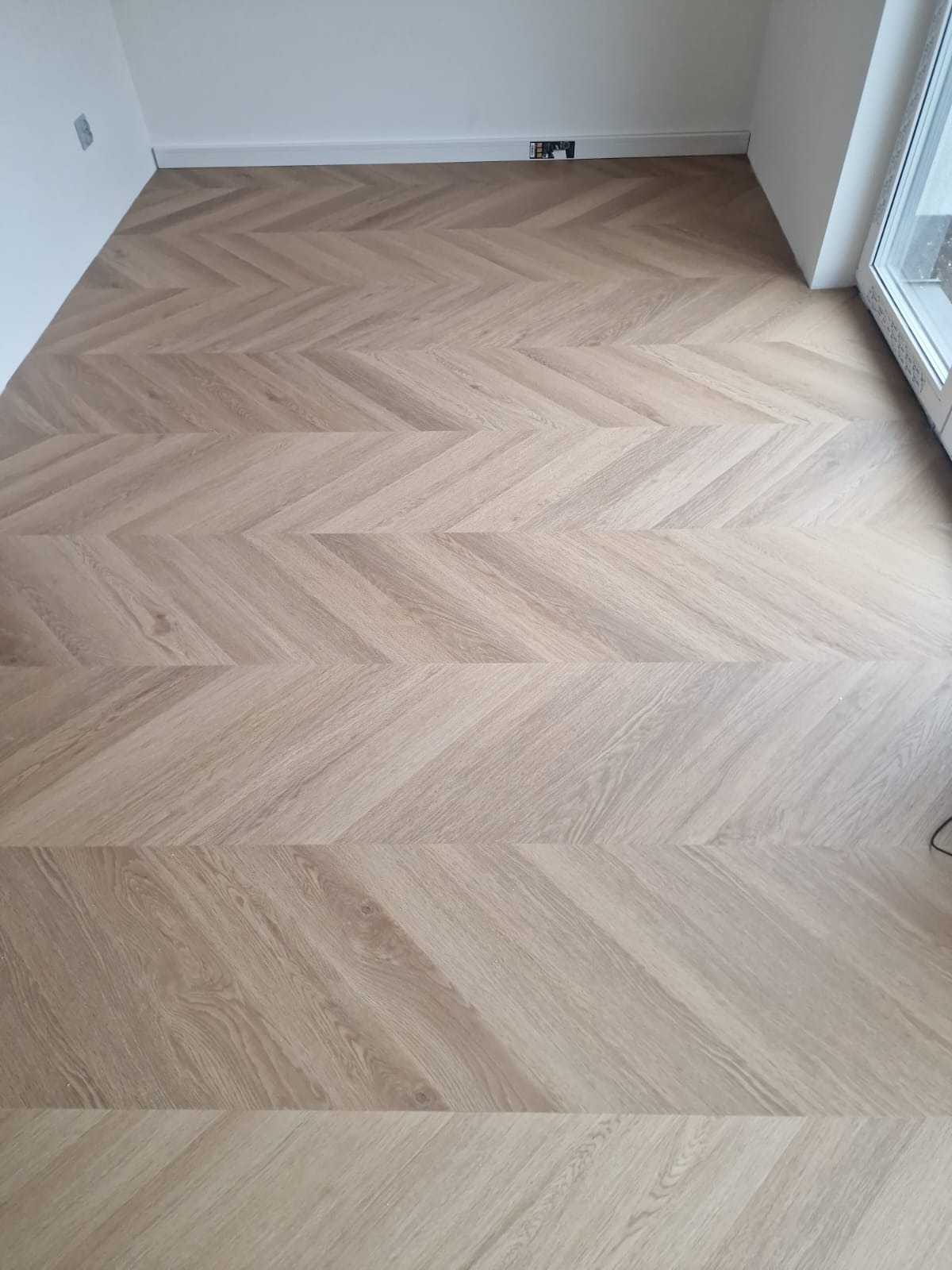 Jasny parkiet ułożony we wzór jodełki francuskiej w narożnym pokoju z białymi ścianami i listwami przypodłogowymi.