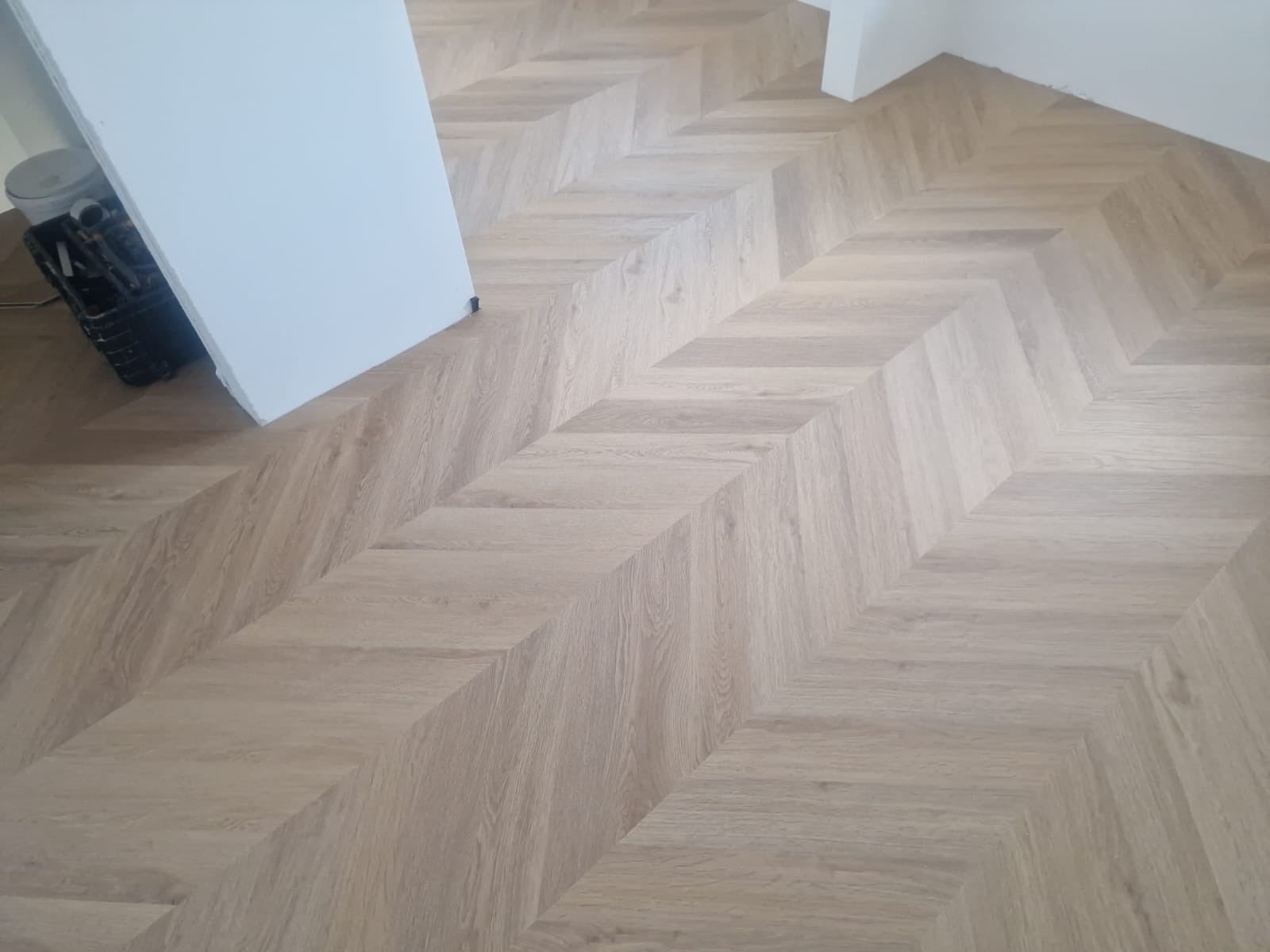 Jasny parkiet ułożony we wzór jodełki francuskiej, widoczny fragment ściany i kosz z narzędziami w rogu.