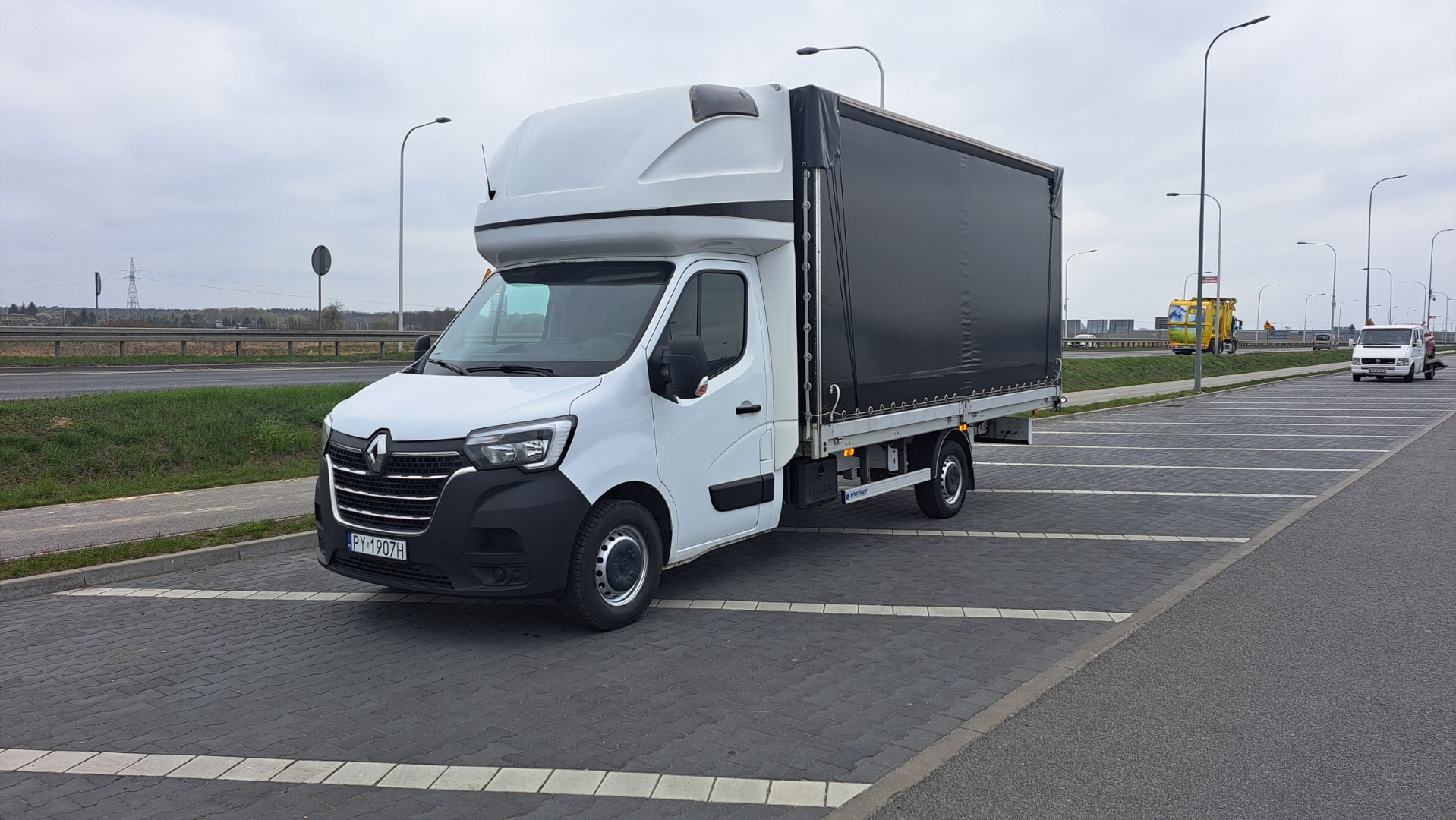 Biały samochód dostawczy Renault Master z plandeką na parkingu z kostki brukowej, widok z przodu, tablica rejestracyjna PY 1907H.