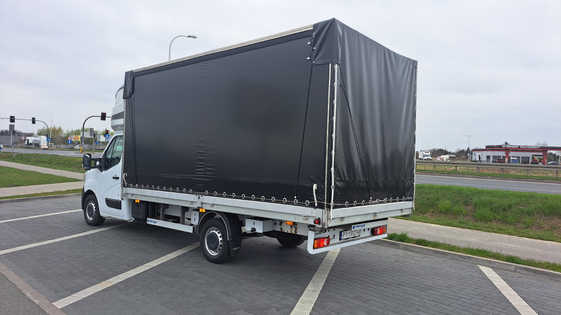 Biały samochód dostawczy z czarną plandeką zaparkowany na parkingu z szarej kostki brukowej, widok z tyłu i boku, w tle stacja benzynowa.