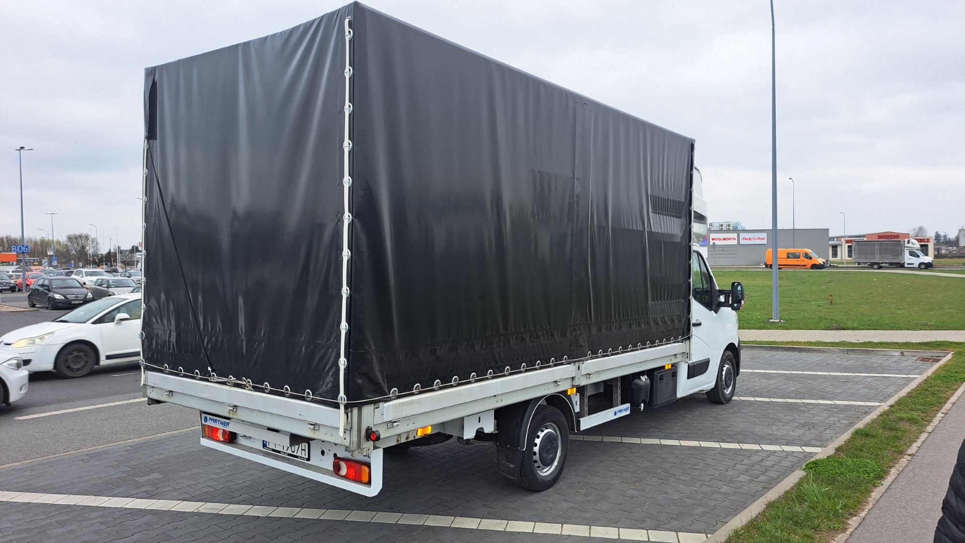 Tył białego samochodu dostawczego z czarną plandeką na parkingu przed budynkiem handlowym w pochmurny dzień.