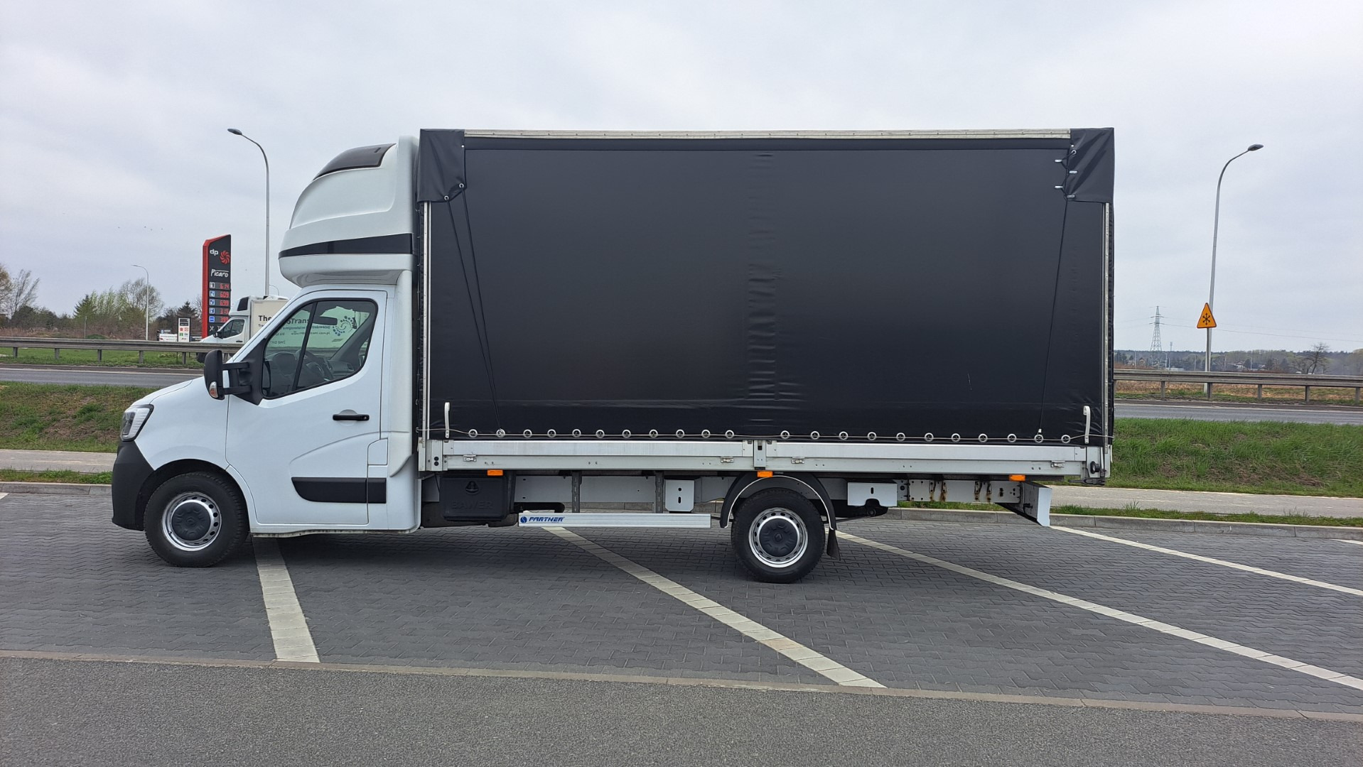 Biały samochód dostawczy z czarną plandeką zaparkowany na parkingu z kostki, widoczny dystrybutor paliwa w tle.