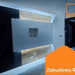 RemFix - Nowoczesna zabudowa RTV z podświetleniem LED, drewnianymi panelami i szarą ścianką z otworem na telewizor. Widoczne gniazdka elektryczne i kable.