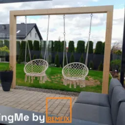 Dwa wiszące fotele hamakowe na drewnianej konstrukcji, widok z zewnątrz, sofa ogrodowa w kadrze, kostka brukowa na pierwszym planie.