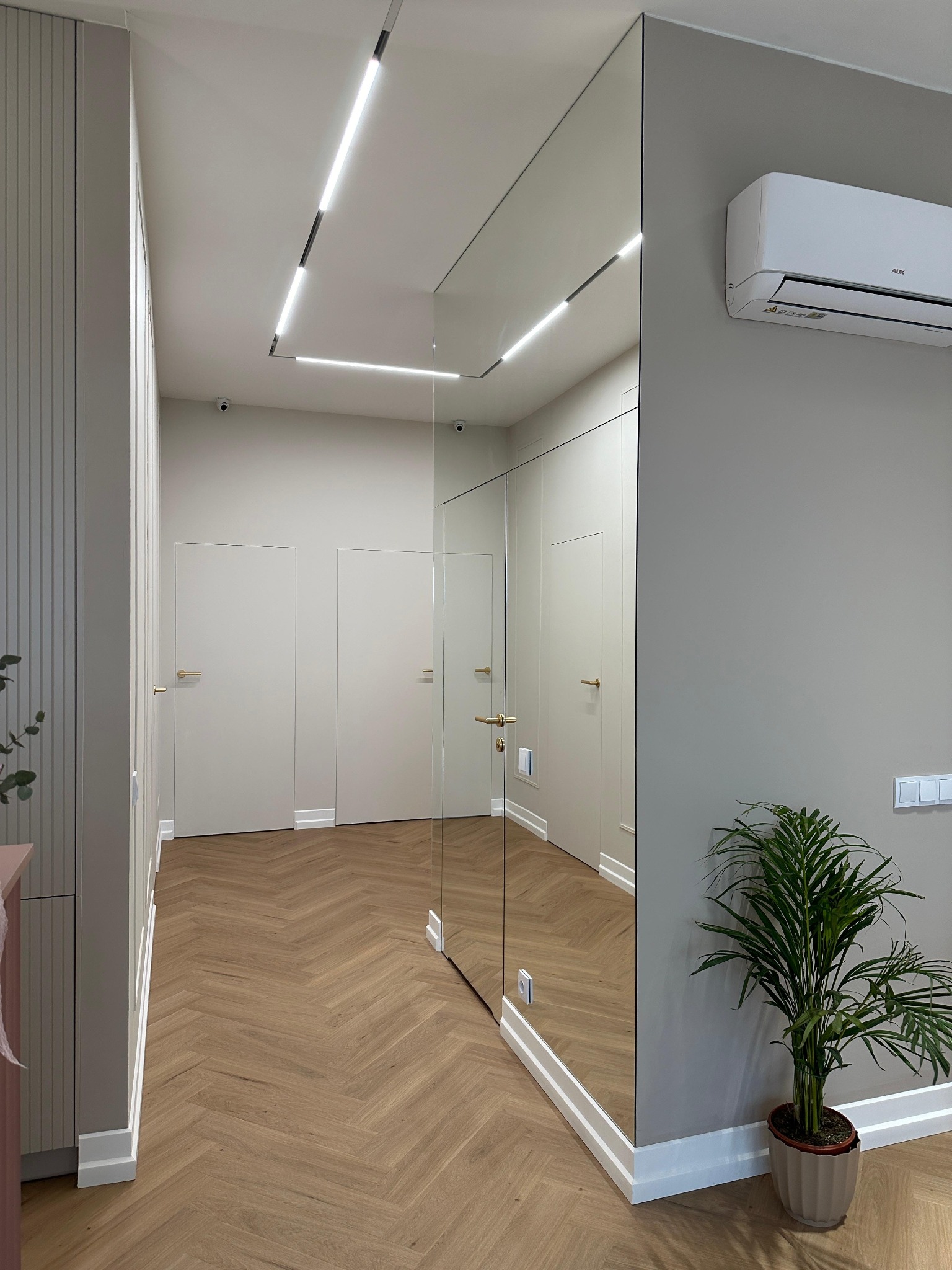 Nowoczesny przedpokój z lustrzaną ścianą, parkietem w jodełkę, minimalistycznym oświetleniem liniowym i klimatyzatorem na szarej ścianie.