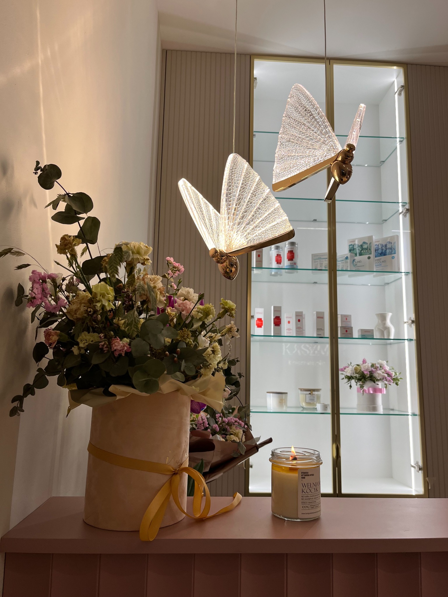 Dekoracja wnętrza salonu kosmetycznego: bukiet kwiatów w pudrowym pudełku, zapalona świeca sojowa oraz dwie wiszące lampy w kształcie motyli na tle przeszklonej witryny z kosmetykami.