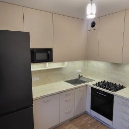 Ex-Mebel Marek Szpak - Jasna kuchnia z beżowymi szafkami, czarną lodówką i piekarnikiem, oraz płytą gazową. Widoczne butelki z wodą przy oknie i fragment osoby w tle.