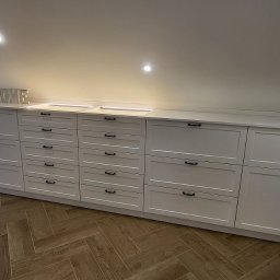 Ex-Mebel Marek Szpak - Białe szafki i szuflady z czarnymi uchwytami, zabudowane pod skosem na poddaszu, z drewnianą podłogą w jodełkę.