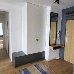 Ex-Mebel Marek Szpak - Wnętrze przedpokoju z zabudowaną szafą wnękową w kolorze białym z ciemnoszarym dołem, oświetleniem punktowym na suficie i drewnianą podłogą zabezpieczoną kartonem.