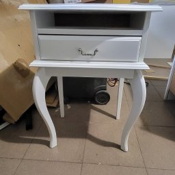 Ex-Mebel Marek Szpak - Biała szafka nocna z szufladą i giętymi nogami, sfotografowana w warsztacie w Lublinie, z widocznymi materiałami stolarskimi w tle.