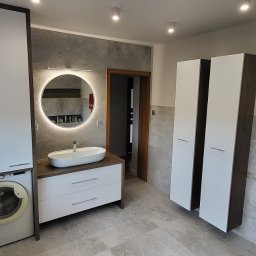 Ex-Mebel Marek Szpak - Nowoczesna łazienka z pralką zabudowaną w szafce, okrągłym lustrem z podświetleniem LED, umywalką nablatową, grzejnikiem drabinkowym z ręcznikami i wysokimi szafkami.