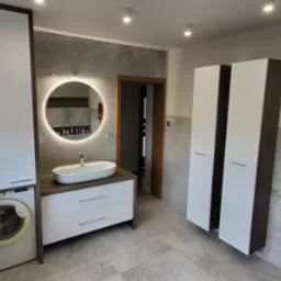 Nowoczesna łazienka z pralką zabudowaną w szafce, okrągłym lustrem z podświetleniem LED, umywalką nablatową, grzejnikiem drabinkowym z ręcznikami i wysokimi szafkami.