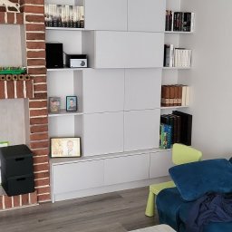 Ex-Mebel Marek Szpak - Biała zabudowa meblowa na ścianie z półkami na książki, elementem dekoracyjnym z cegły i widocznym fragmentem salonu z niebieską kanapą oraz stolikiem kawowym z pilotem i kubkiem.