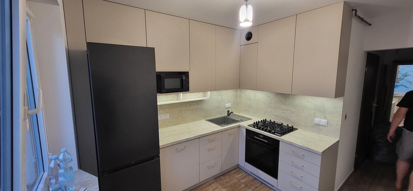 Jasna kuchnia z beżowymi szafkami, czarną lodówką i piekarnikiem, oraz płytą gazową. Widoczne butelki z wodą przy oknie i fragment osoby w tle.