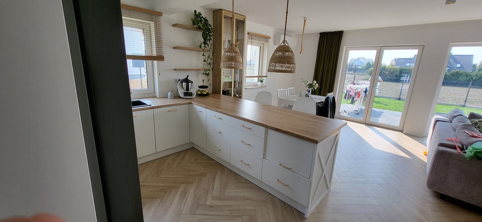 Jasna kuchnia z drewnianym blatem, wyspą z szufladami i złotymi uchwytami, podłoga ułożona w jodełkę, widok na ogród przez duże okno.